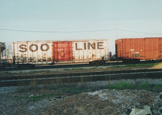 Two freight cars, one white with 'SOO LINE' lettering and the other orange-red with 'SOU' markings, are parked on adjacent tracks in a gravel yard.