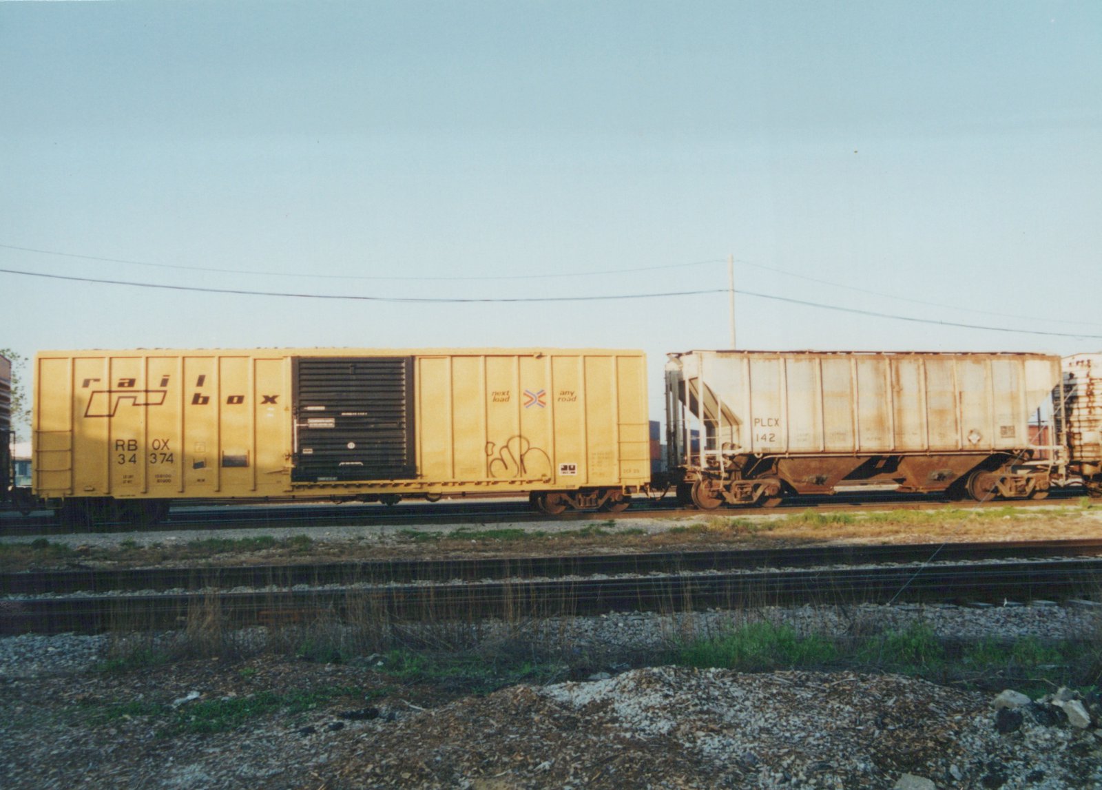 A side view of two freight cars, a yellow Railbox car and a white PLCX car, on a set of railroad tracks with a clear blue sky above.