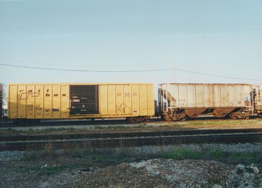 A side view of two freight cars, a yellow Railbox car and a white PLCX car, on a set of railroad tracks with a clear blue sky above.