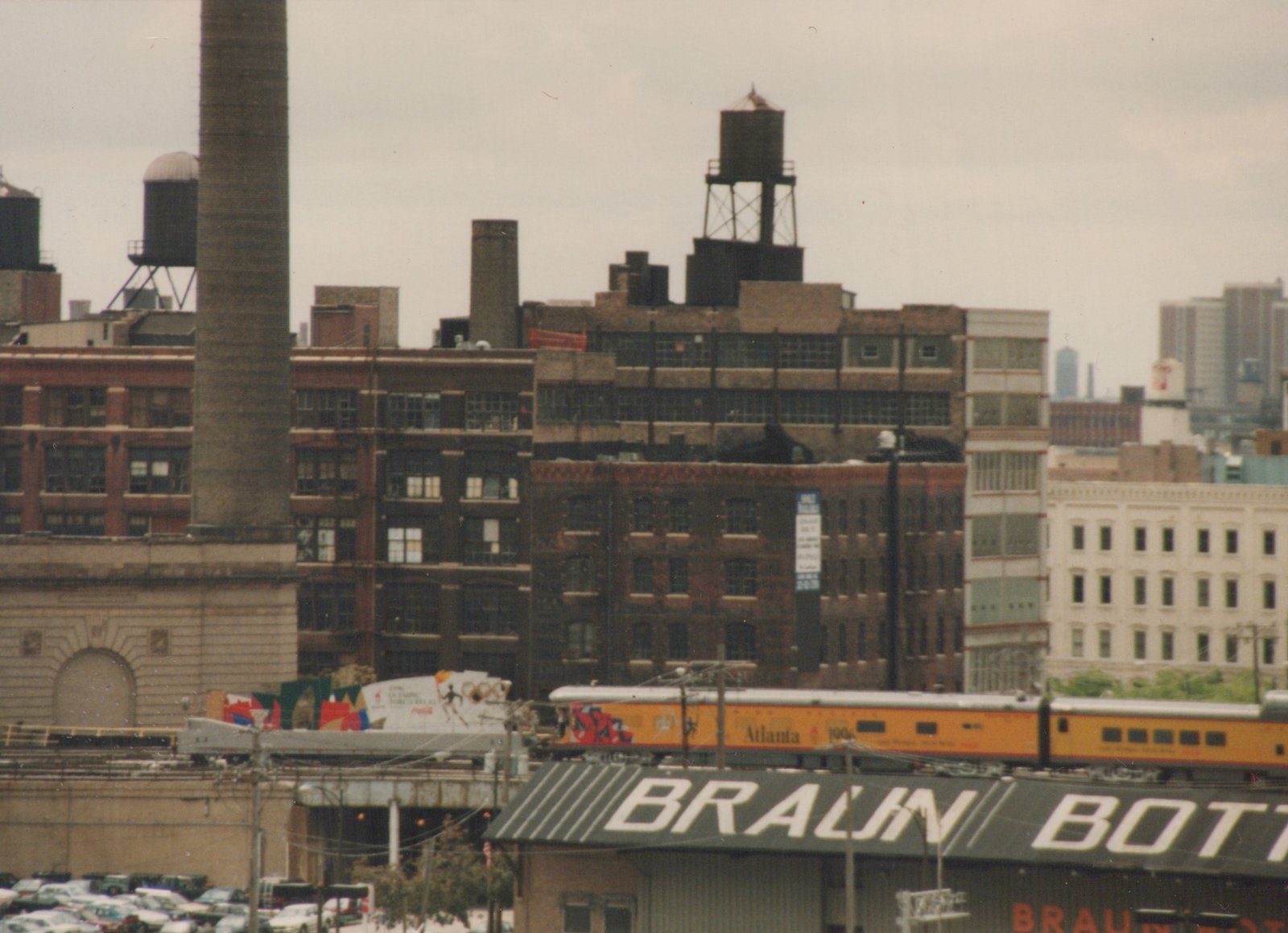 An orange and silver double-decker commuter train with 'Atlanta 1996' on its side travels on an elevated track in a city with brick buildings and a large chimney in the background.