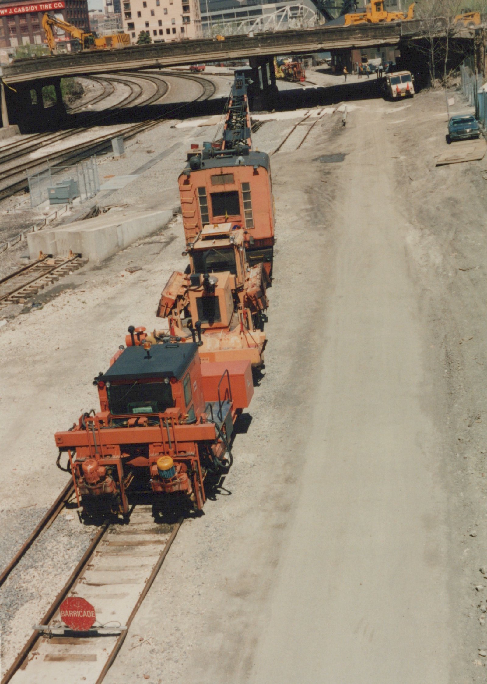 Three orange track maintenance vehicles on a set of train tracks under an elevated highway, with construction equipment and a few people in the background.