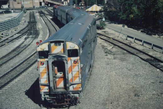 A high-angle view of a double-decker Metra train car, silver with orange and white striped ends, stopped on tracks in a gravel yard on a sunny day.