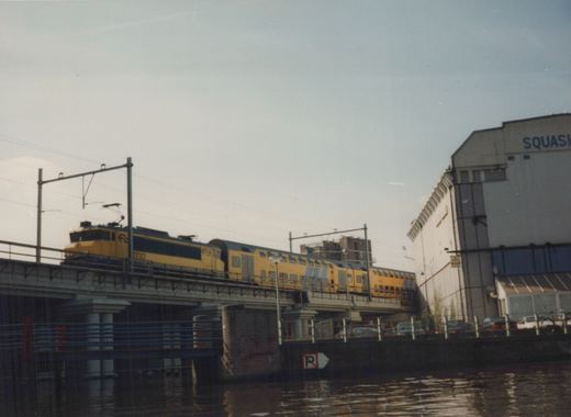A yellow and blue electric train, led by locomotive 1732, travels on an elevated bridge over water, with a building labeled 'SQUASH' and parked cars nearby.