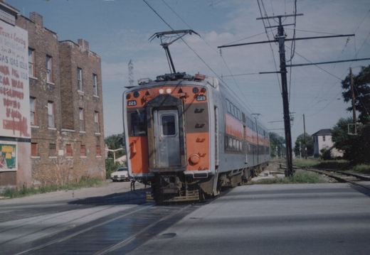 A silver and orange bi-level electric train with the number 225 is stopped on tracks that cross a street in a city, with a brick building and a billboard in the background.