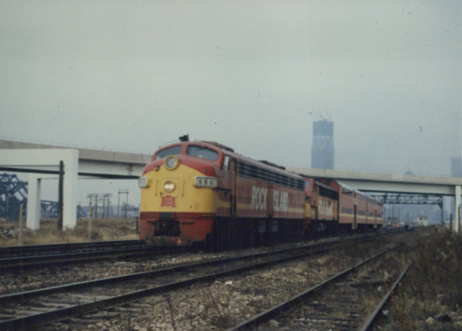 A red and yellow Rock Island F7 locomotive numbered 660 pulls a line of passenger cars on railroad tracks under an overpass, with a tall skyscraper visible in the hazy background.