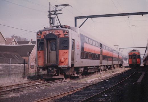 A silver and orange double-decker commuter train car with the number 1548 on its side, parked in a rail yard next to another identical car with number 345, under an overcast sky. The logo on the side is a red 'I'.