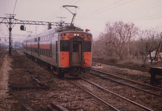 A silver and orange double-decker commuter train with the number 810 is on a set of tracks, with bare trees and overcast sky in the background.
