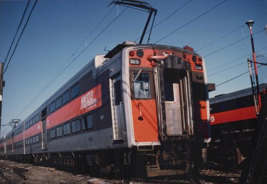 A red, gray, and black Metra Highliner train car, number 505, shown from the front three-quarter view on a sunny day, with another car and overhead power lines in the background.