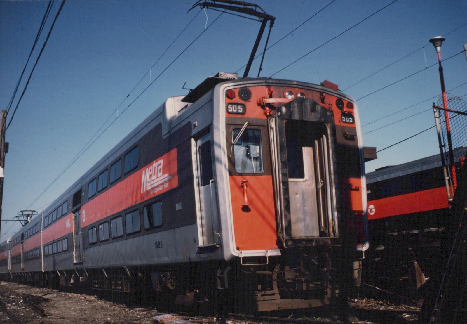 A red, gray, and black Metra Highliner train car, number 505, shown from the front three-quarter view on a sunny day, with another car and overhead power lines in the background.