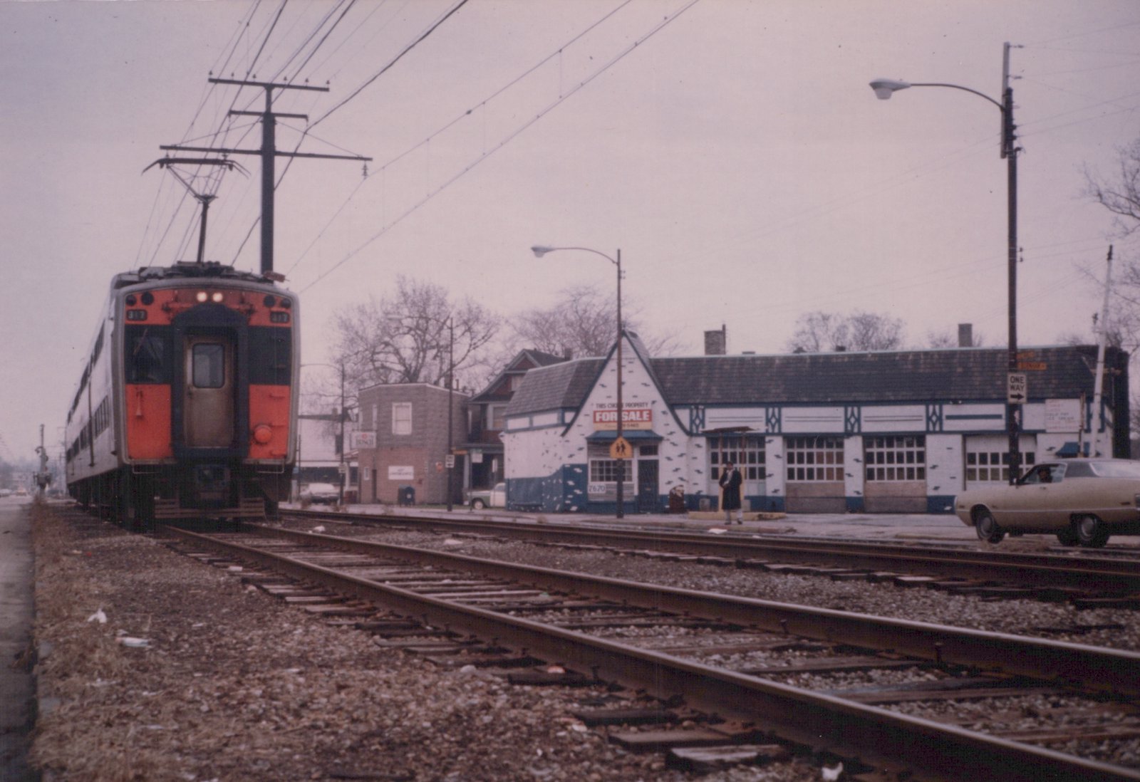 A silver and orange electric train car, number 317, is stopped at a station platform next to a white building with a 'For Sale' sign, under an overcast sky in 1978.