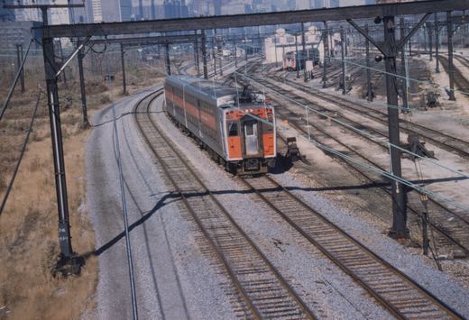 A silver and orange double-decker electric train car with the number 119 on its front moves along curved tracks in a sunlit rail yard with multiple overhead wires and a city skyline in the background.