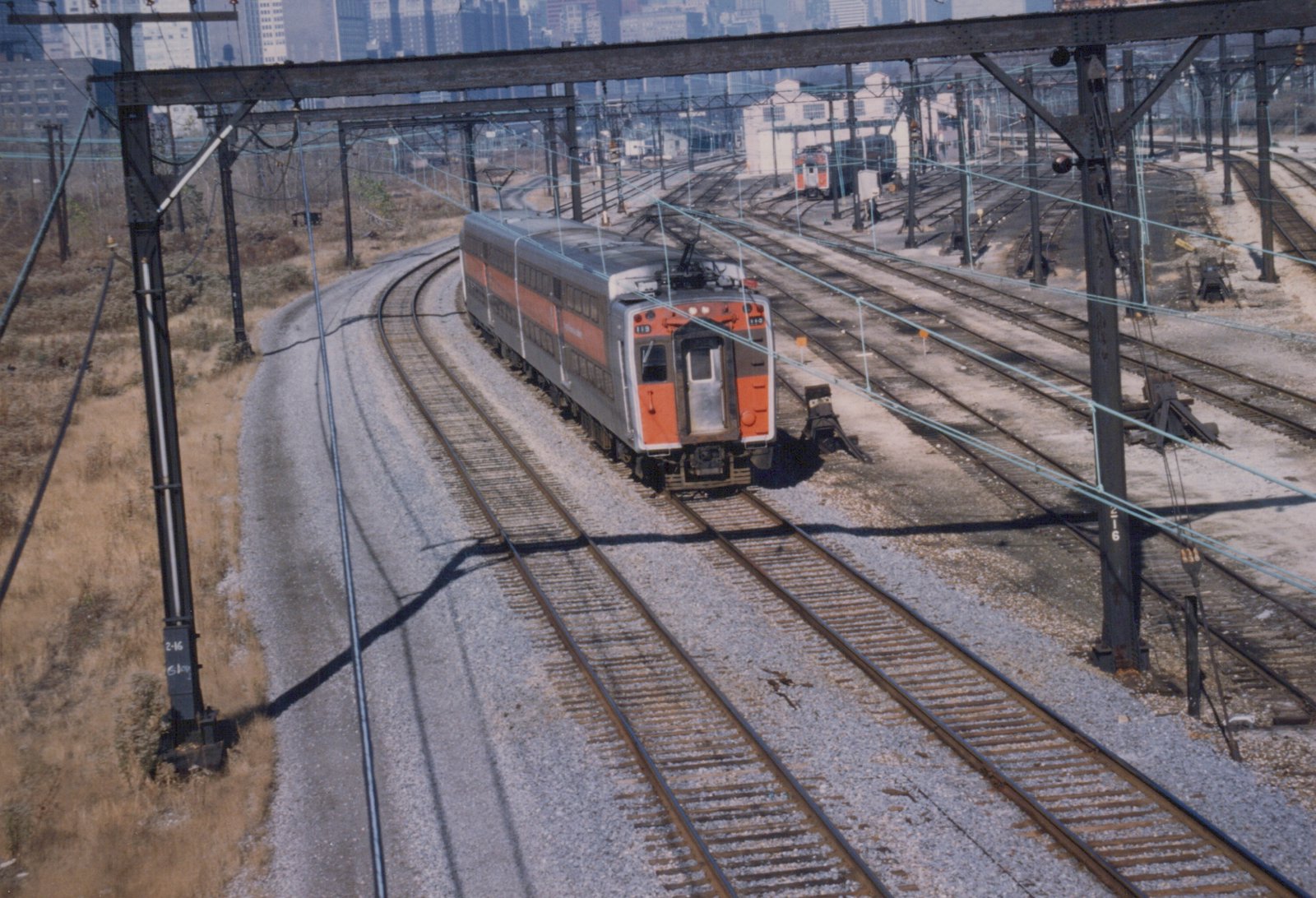 A silver and orange double-decker electric train car with the number 119 on its front moves along curved tracks in a sunlit rail yard with multiple overhead wires and a city skyline in the background.