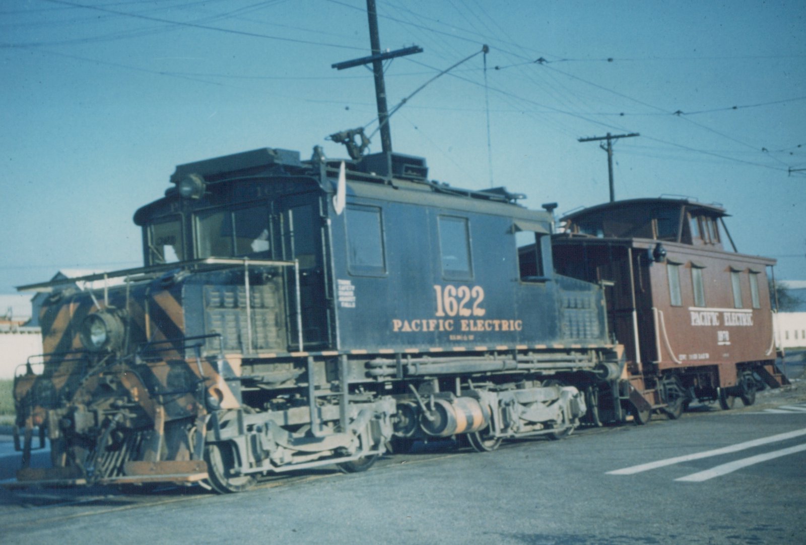 A dark blue Pacific Electric Railway electric locomotive numbered 1622 is coupled to a brown caboose on a street with overhead wires.