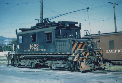 A dark green Pacific Electric electric locomotive numbered 1622, with a yellow and black striped front, is parked on a street under overhead wires, with a tan passenger car partially visible behind it.