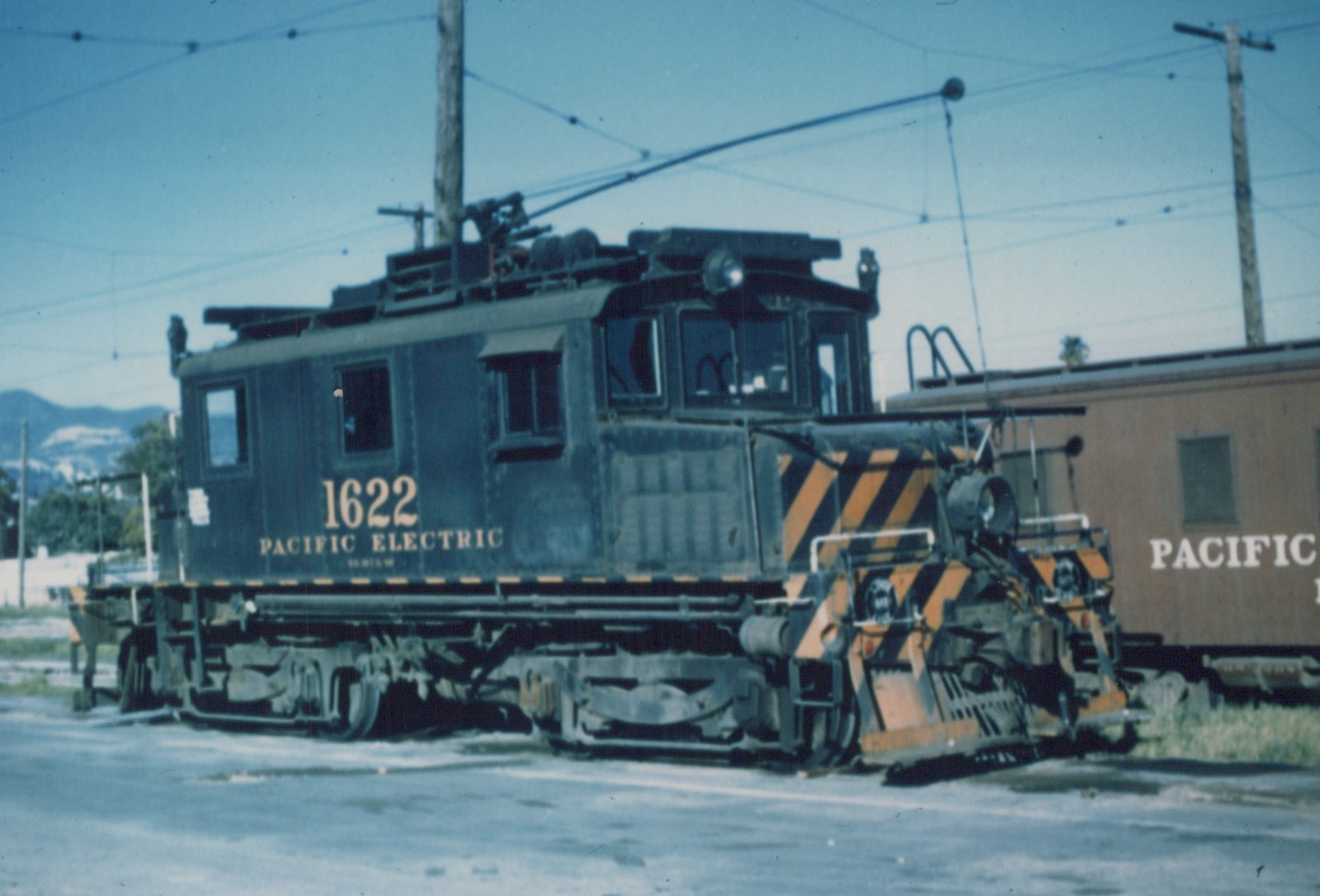 A dark green Pacific Electric electric locomotive numbered 1622, with a yellow and black striped front, is parked on a street under overhead wires, with a tan passenger car partially visible behind it.