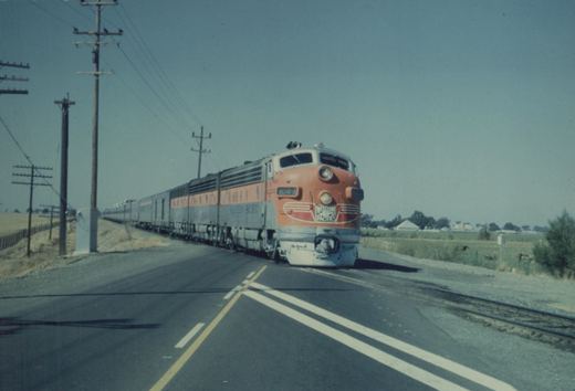 A red and silver Western Pacific diesel locomotive numbered 804-A is crossing a paved road, pulling a long line of passenger cars through a flat, rural landscape.