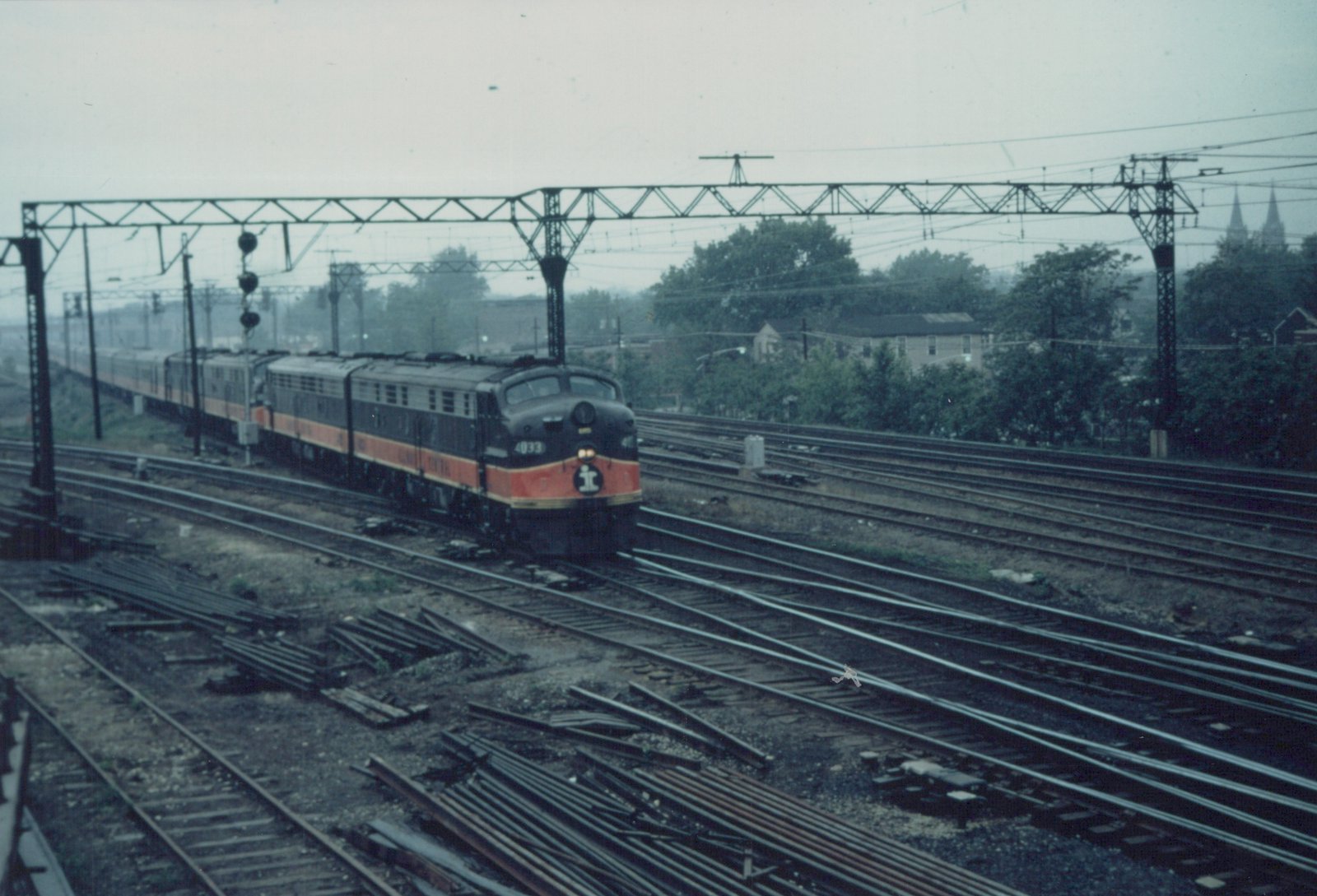 An Illinois Central train with a black and orange E8 locomotive pulling passenger cars on a multi-track rail yard with overhead wires, under an overcast sky.