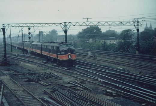 An Illinois Central train with a black and orange E8 locomotive pulling passenger cars on a multi-track rail yard with overhead wires, under an overcast sky.