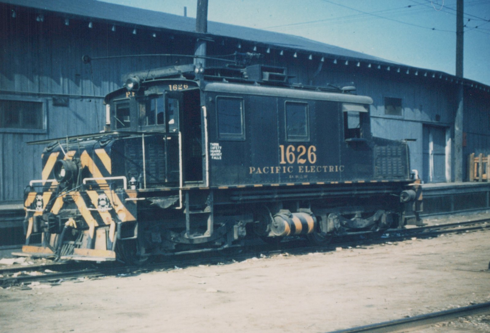 A dark blue Pacific Electric Railway electric locomotive number 1626, with yellow and black safety stripes, sits on a track in a yard next to a large building.