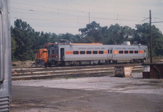 A silver and orange passenger railcar labeled Chicago South Shore and South Bend Railroad, number 10, sits on tracks in a gravel yard with overhead wires and trees in the background.