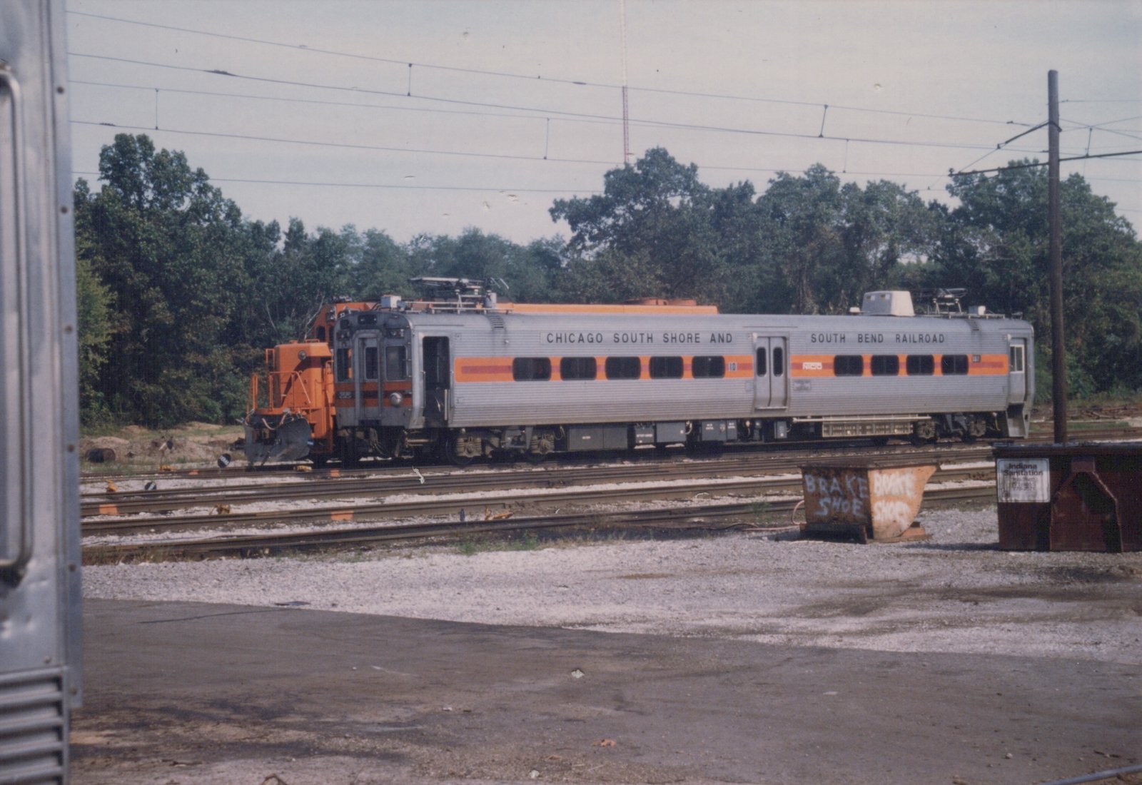 A silver and orange passenger railcar labeled Chicago South Shore and South Bend Railroad, number 10, sits on tracks in a gravel yard with overhead wires and trees in the background.
