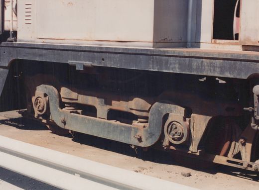 Close-up of the underframe and truck of a rail vehicle, showing a rusty metal frame with the number -4202 painted on it, on a dirt ground beside a concrete curb.