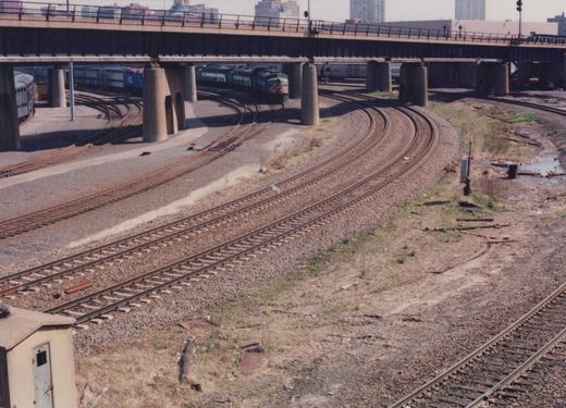A Metra train with a green locomotive and double-decker passenger cars travels under a large concrete bridge on a curved track in a gravel yard, with city buildings in the distance.