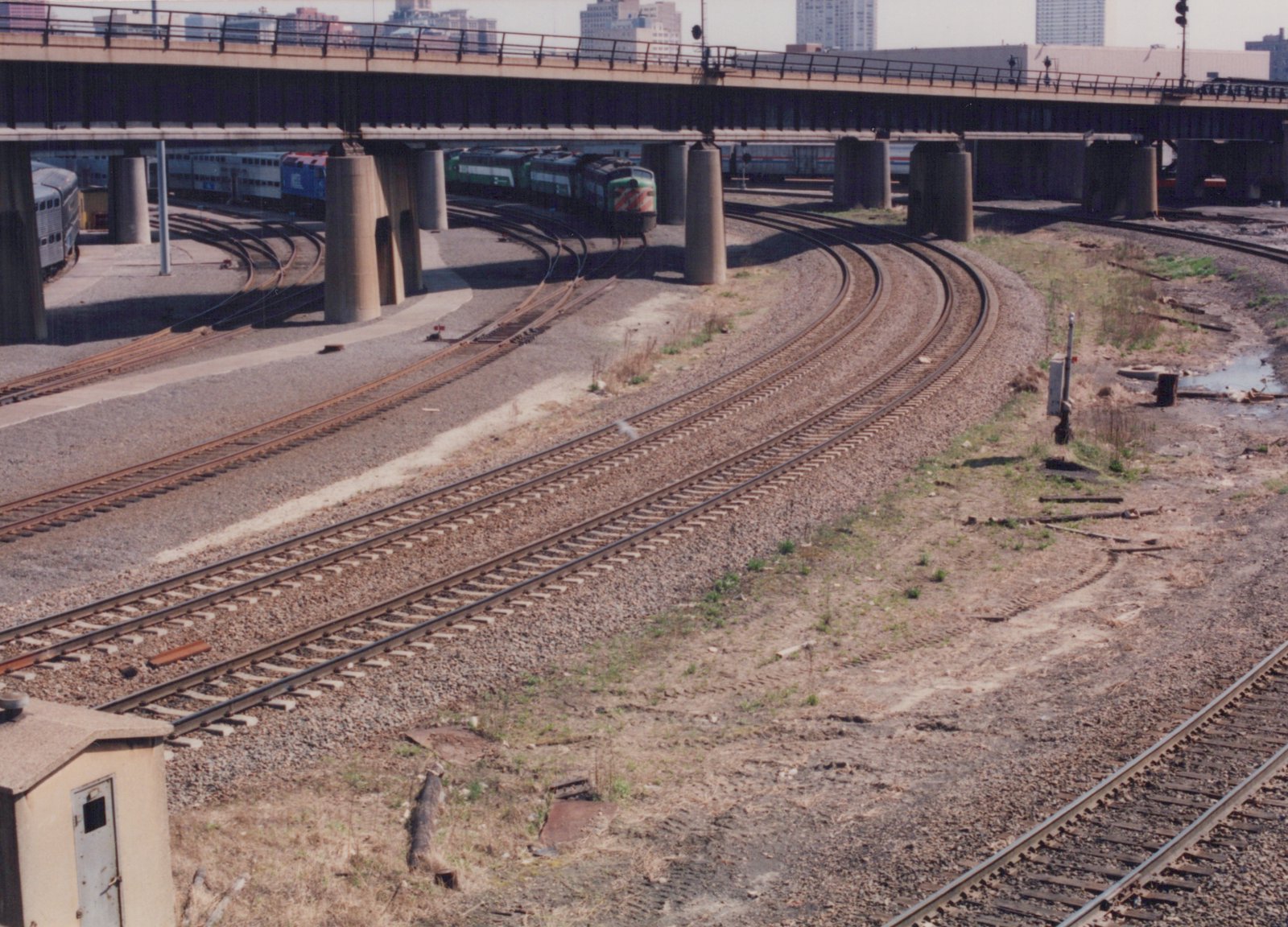 A Metra train with a green locomotive and double-decker passenger cars travels under a large concrete bridge on a curved track in a gravel yard, with city buildings in the distance.