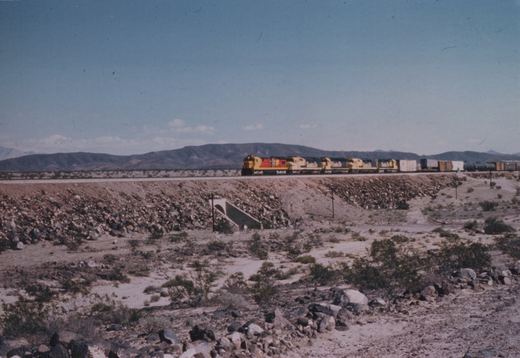 A long freight train with yellow and red locomotives moves along a track through a barren, rocky desert with distant mountains under a clear sky.