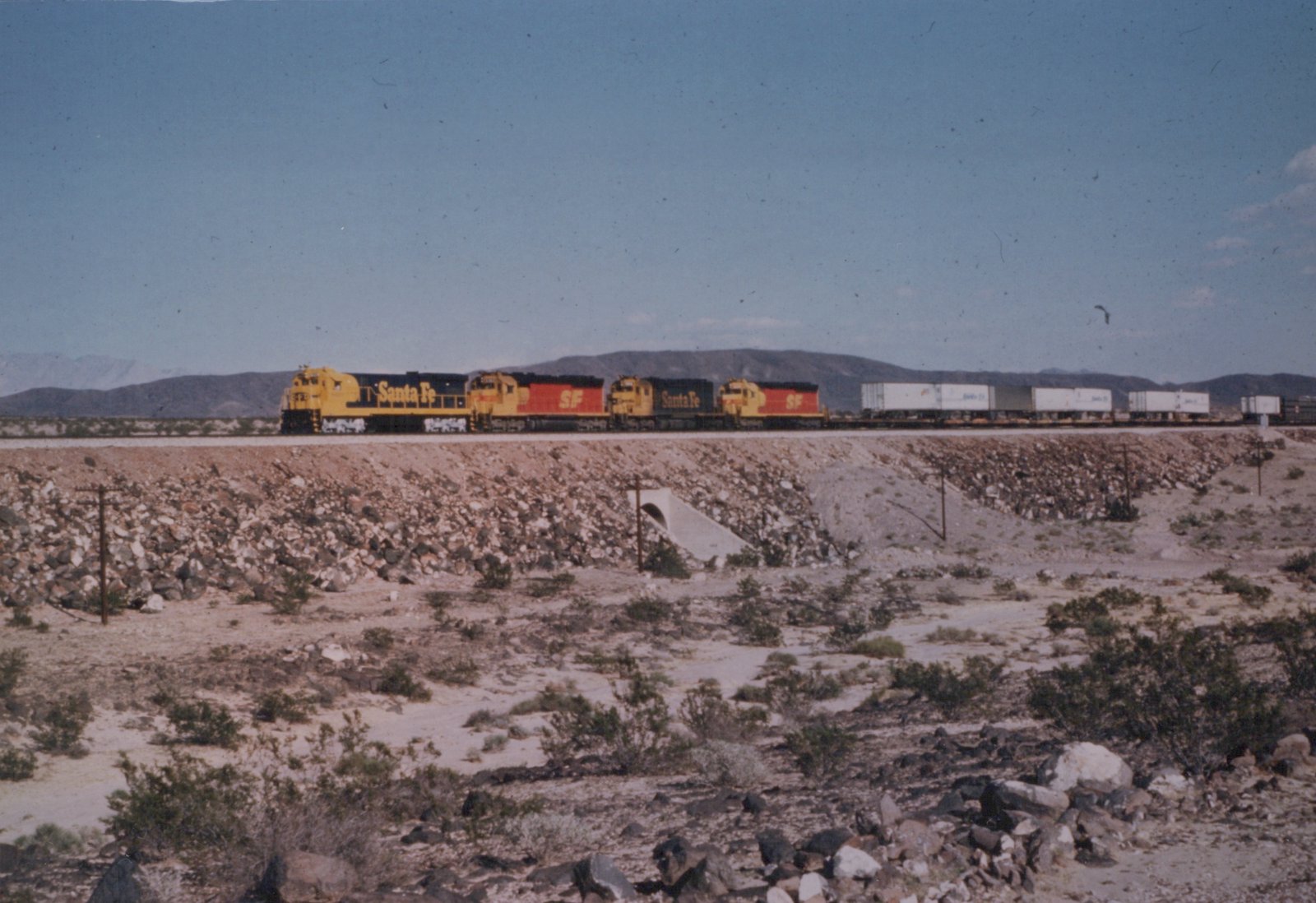 A side view of a Santa Fe freight train moving across a rocky, arid landscape under a clear blue sky.