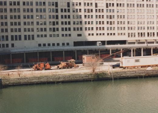A photo of a rail yard beside a river, featuring orange maintenance machines, freight cars, and vehicles, with a large building and elevated road in the background.