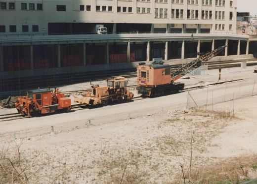 A large orange rail crane and two other pieces of orange track maintenance machinery are parked on railway tracks next to a large, multi-story building with an elevated roadway.