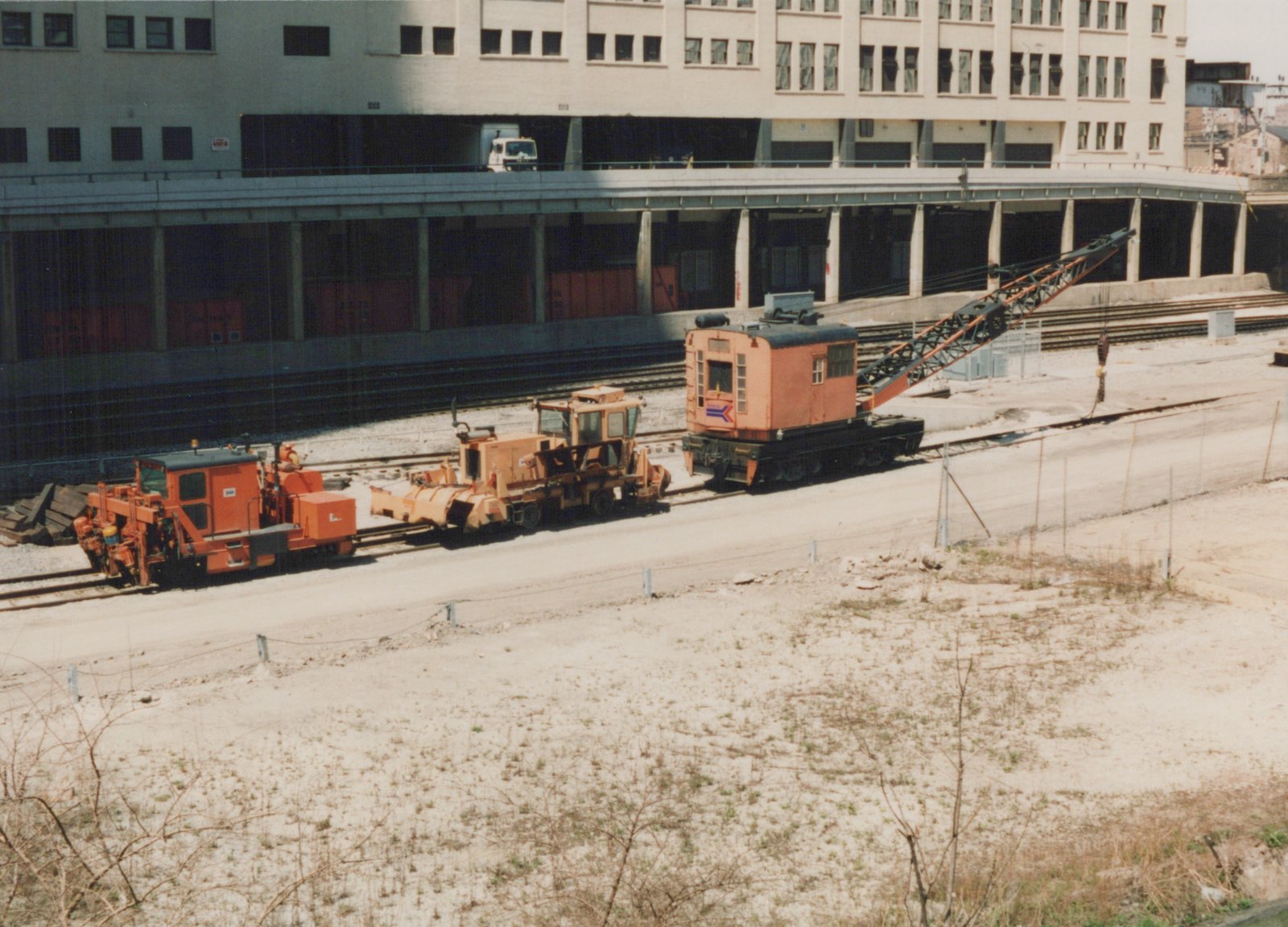 A large orange rail crane and two other pieces of orange track maintenance machinery are parked on railway tracks next to a large, multi-story building with an elevated roadway.