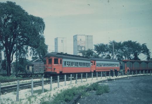 Two red and gray interurban cars on a railway track in a wooded area, with a large grain elevator in the background.