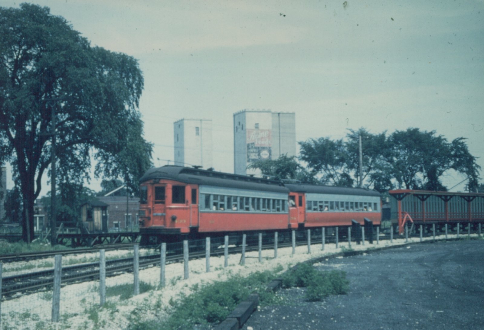 Two red and gray interurban cars on a railway track in a wooded area, with a large grain elevator in the background.