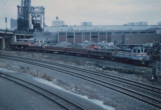 An Amtrak locomotive in white, red, and blue livery pulls a train of open-top freight cars through a gravel yard on an overcast winter day, with an industrial building and a drawbridge in the background.
