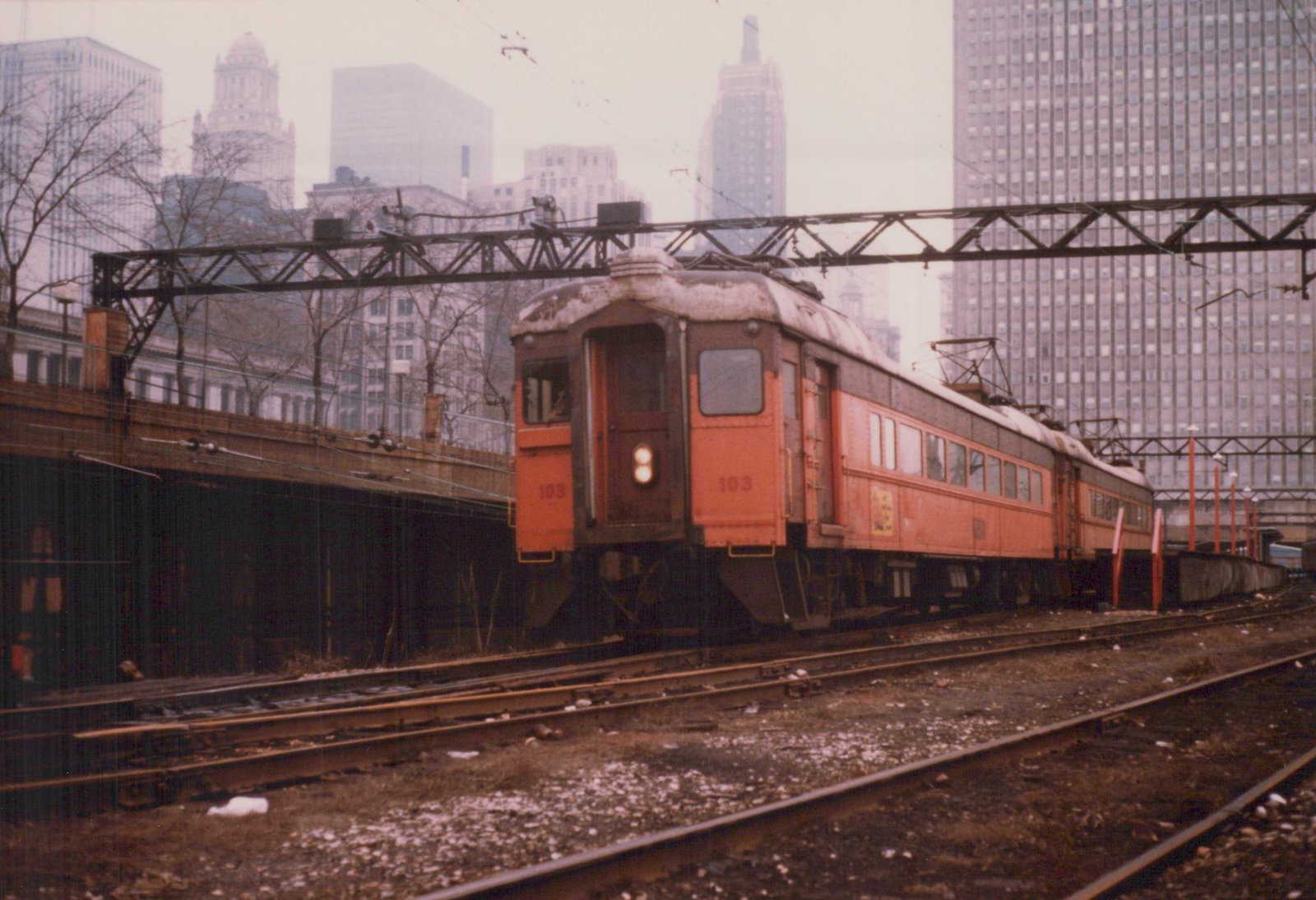 An orange and dark brown interurban train on elevated tracks with snow on the ground and a city skyline in the background.