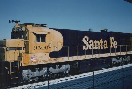 A side view of a Santa Fe diesel locomotive numbered 9506, painted in yellow and dark blue, standing on a track next to a chain-link fence under a clear blue sky.