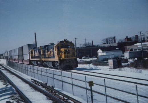 A yellow and black Santa Fe diesel locomotive numbered 9542 pulls a long freight train of double-stacked containers on a snow-covered track next to a chain-link fence.