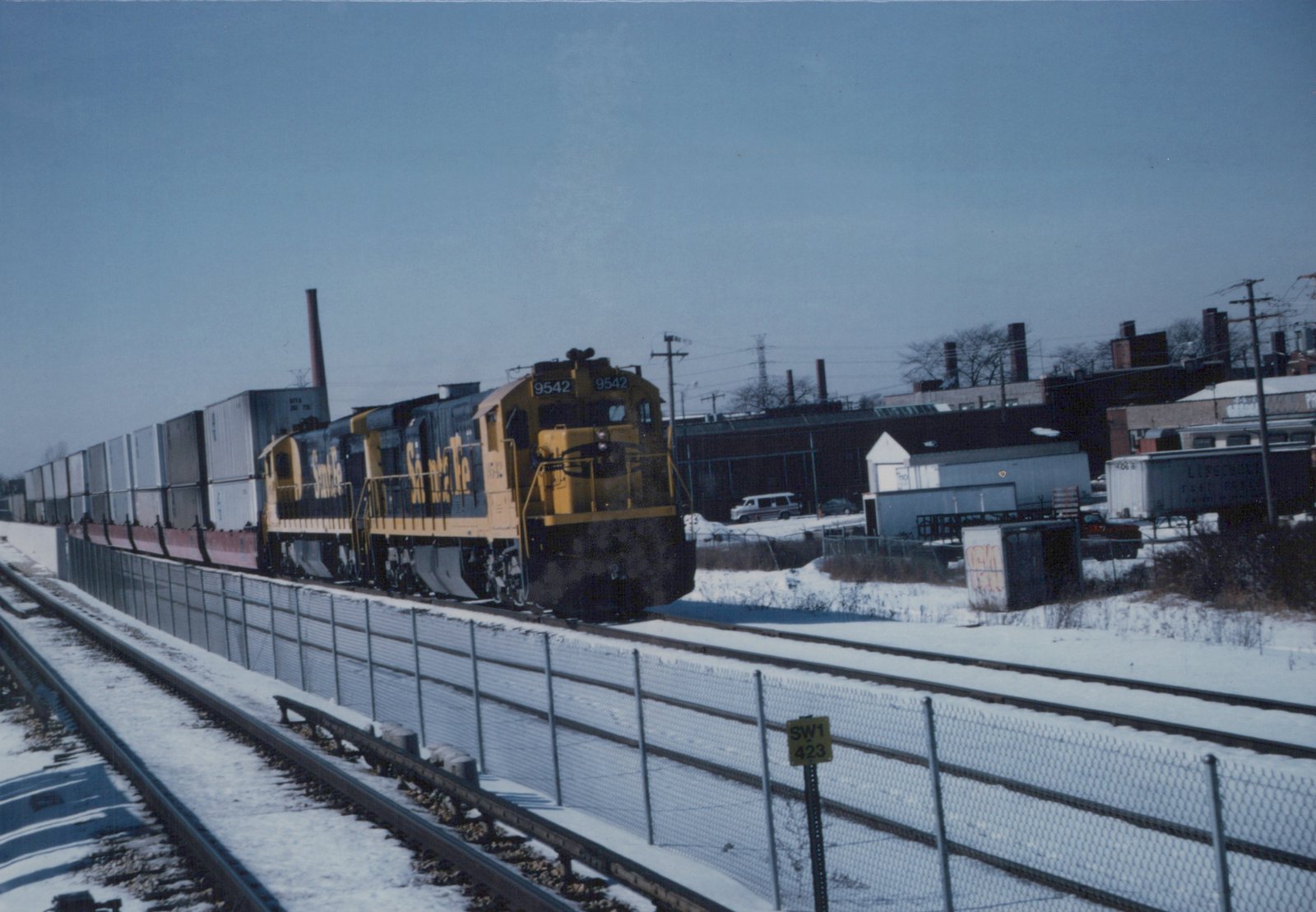 A yellow and black Santa Fe diesel locomotive numbered 9542 pulls a long freight train of double-stacked containers on a snow-covered track next to a chain-link fence.