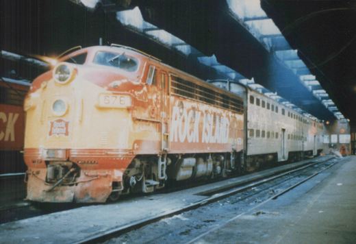 A yellow and orange Rock Island diesel locomotive, number 676, is at a station platform pulling silver passenger cars under an arched roof with skylights.
