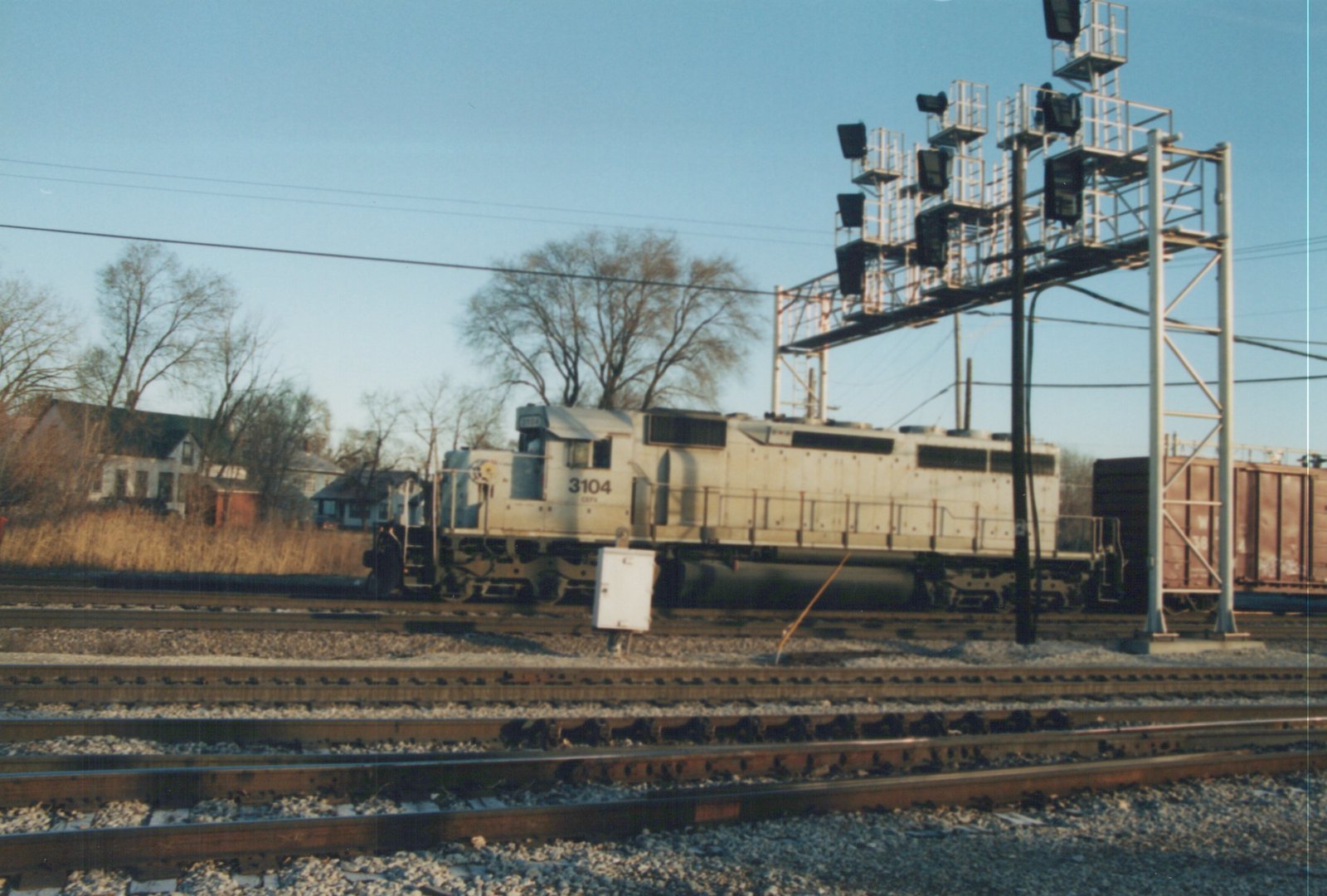 A silver diesel locomotive numbered 3104 with CEPY reporting marks is parked on railroad tracks next to a large signal gantry, with bare trees and houses in the background.