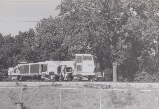 A large white Metra work machine with the number PST 23288 sits on railroad tracks beside a chain-link fence, with dense trees in the background.