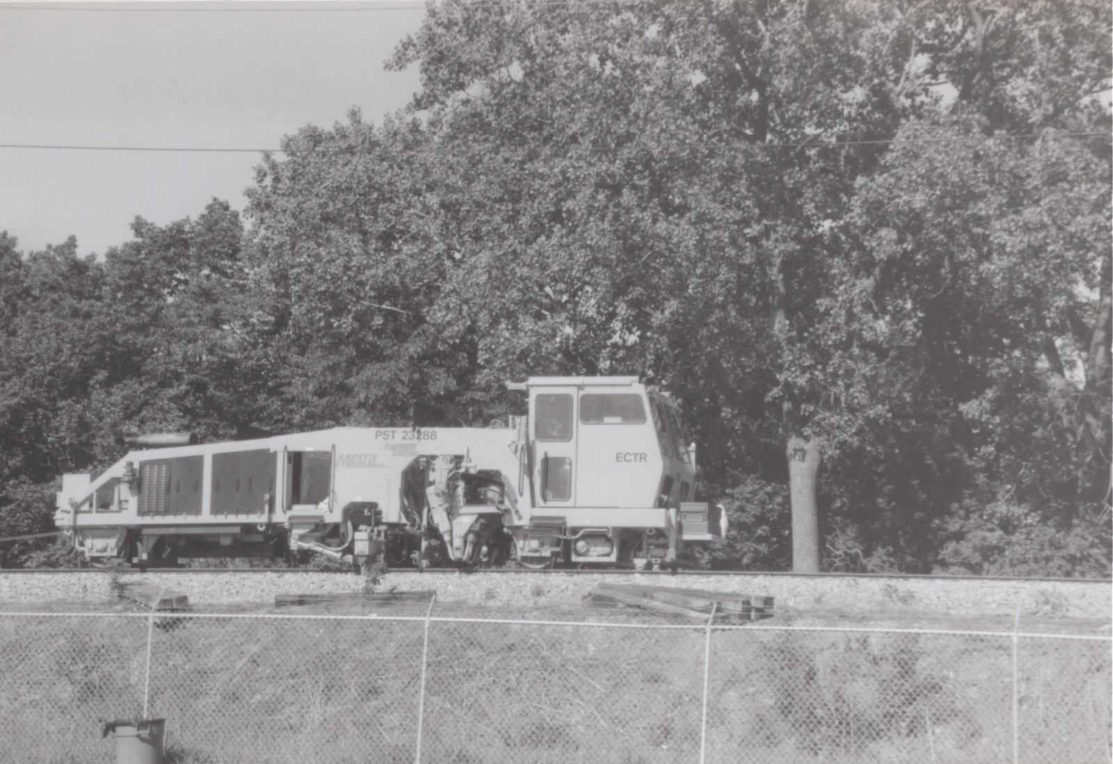 A large white Metra work machine with the number PST 23288 sits on railroad tracks beside a chain-link fence, with dense trees in the background.