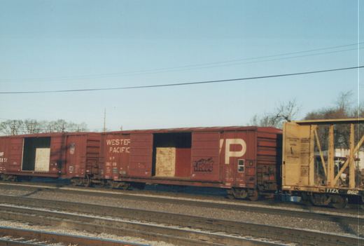 A line of freight cars on a railroad track, including two red Western Pacific boxcars and a yellow TTX flatcar, with bare trees and overhead wires in the background.