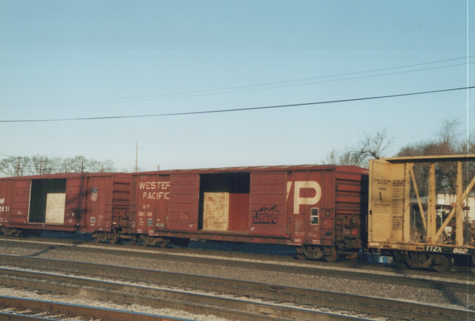 A line of freight cars on a railroad track, including two red Western Pacific boxcars and a yellow TTX flatcar, with bare trees and overhead wires in the background.