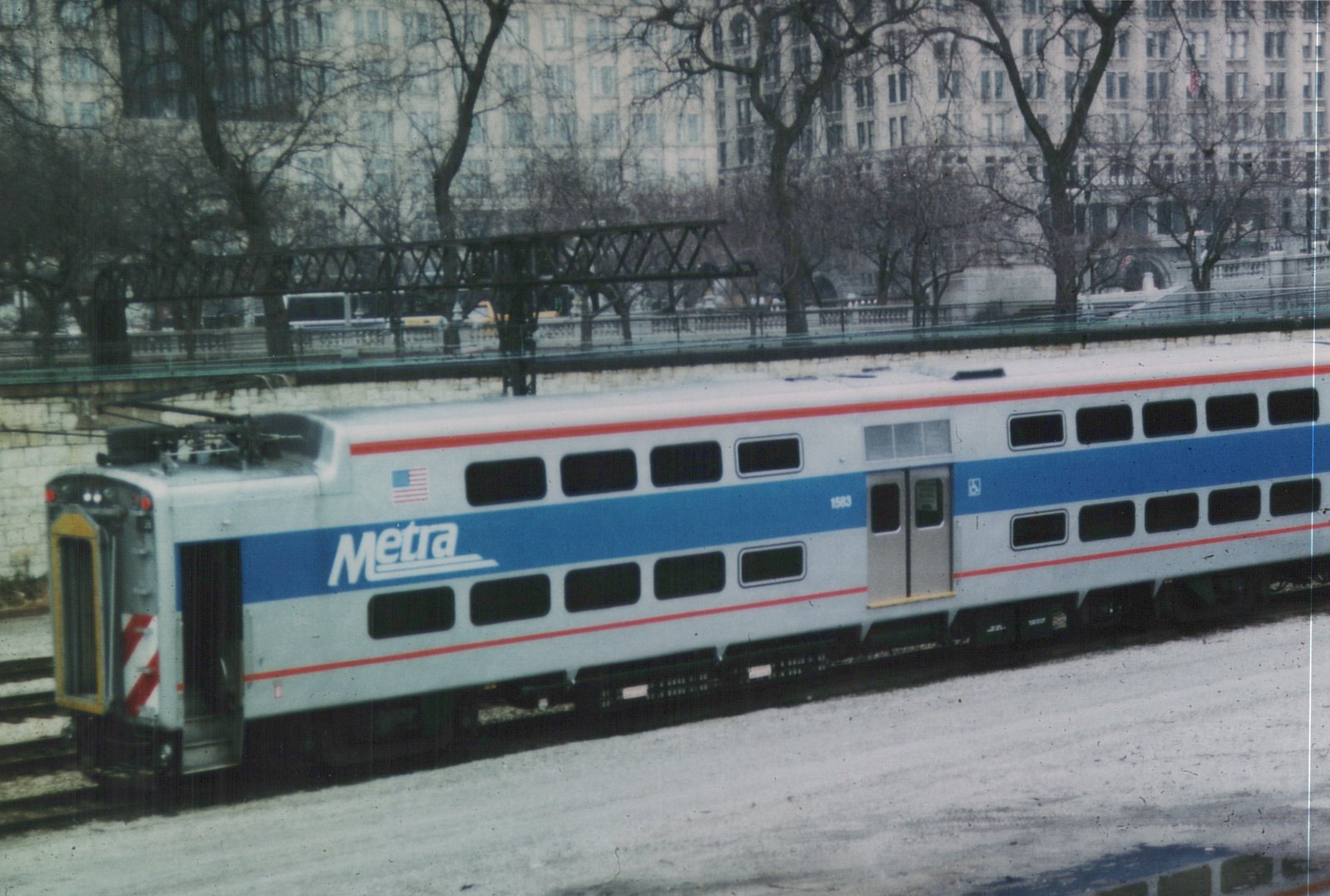 A side view of a silver and blue Metra Highliner train car, number 1583, on a snowy elevated track with a city building in the background.