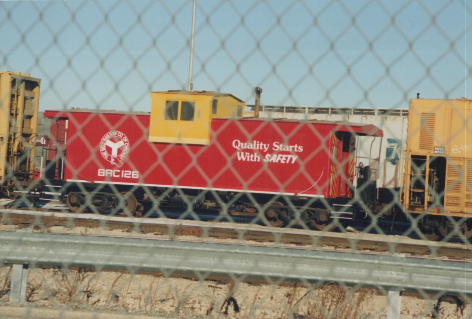 A red and yellow caboose, marked BRC 126 for The Belt Railway Co of Chicago, sits on railroad tracks behind a chain-link fence, with the slogan 'Quality Starts With SAFETY' on its side.