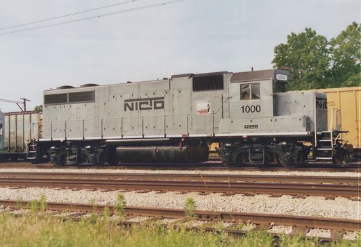 A silver Northern Indiana Commuter Transportation District diesel locomotive numbered 1000 is shown in profile on railroad tracks surrounded by freight cars and greenery, under an overcast sky.