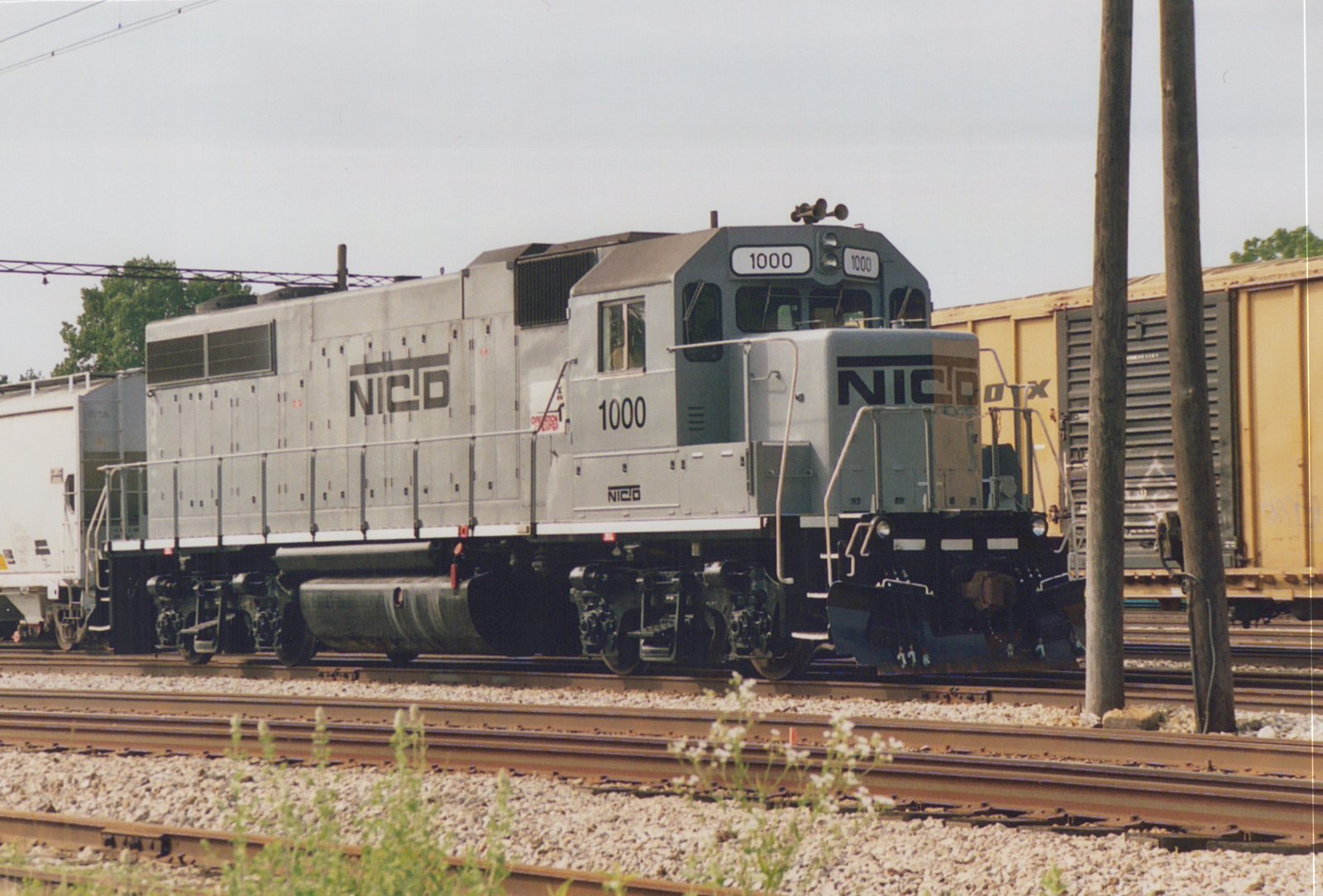 A silver NICO 1000 EMD SW1200 diesel locomotive is parked on railroad tracks in a yard, with yellow and white freight cars nearby under an overcast sky.